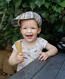 Child wearing a hat and smiling and holding an ice cream cone, with ice cream covering their nose and mouth.
