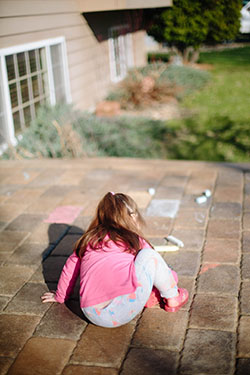 Child with long hair draws with chalk on patio tiles.