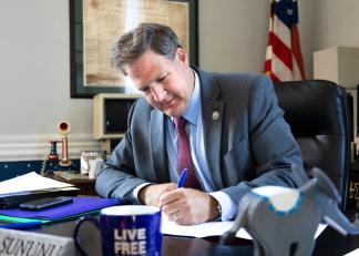 Governor Sununu sitting at a desk and signing a sheet of paper with a blue pen